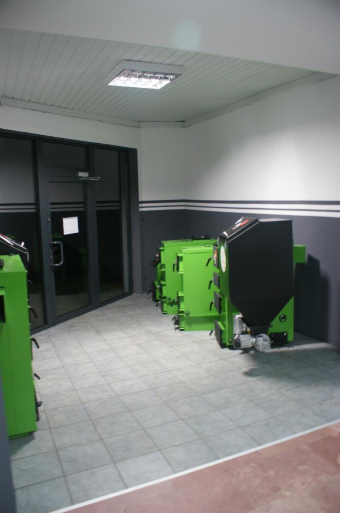 Nowe, zielone piece na paliwo stałe ustawione w rzędzie w pomieszczeniu z szarą podłogą i czarno-białymi ścianami. Widoczny fragment drzwi wejściowych.