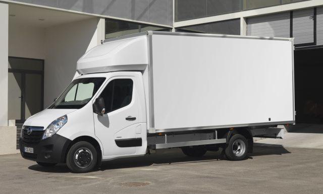 Biały samochód dostawczy Opel Movano z zabudową kontenerową na tle budynku. Widoczny z boku, lekko od przodu. Oświetlenie dzienne, cień pod pojazdem.