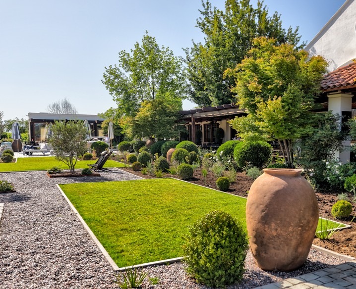 Nowoczesny ogród z trawnikiem, ścieżką żwirową i ozdobnymi krzewami. Duża gliniana donica na pierwszym planie, pergola w tle. Słoneczny dzień.