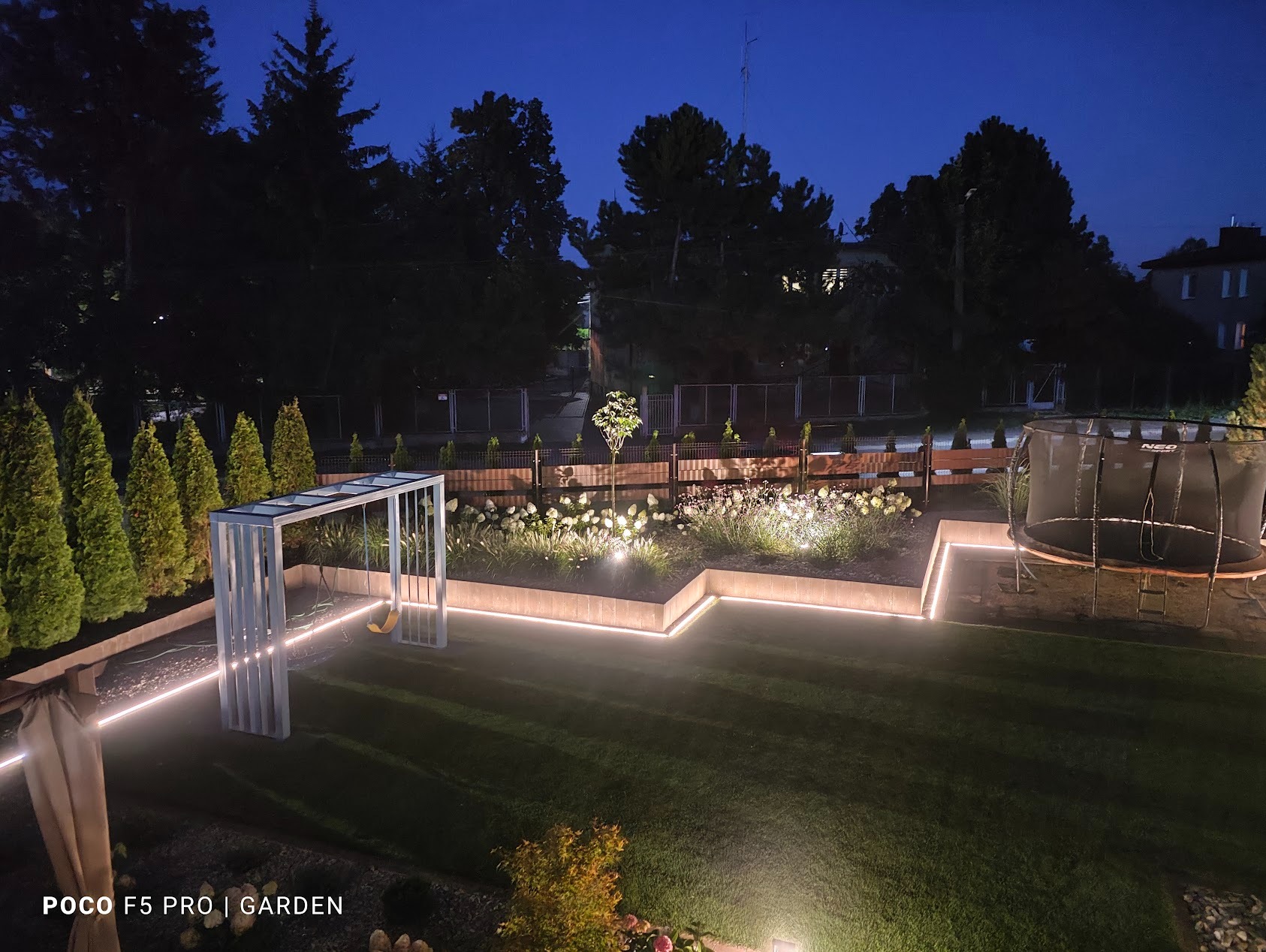 Oświetlony ogród nocą z równo przystrzyżonym trawnikiem, rabatami kwiatowymi i trampoliną. Dekoracyjne oświetlenie LED podkreśla architekturę ogrodu.