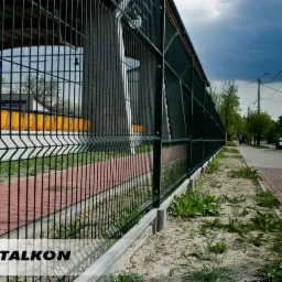 Długi, zielony system ogrodzeniowy z paneli, zamontowany na betonowej podmurówce, oddzielający chodnik z czerwonej kostki od terenu zielonego z chwastami. W tle widoczny budynek i pochmurne niebo.