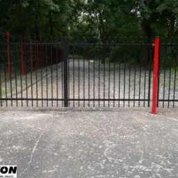 Czarne ogrodzenie panelowe z bramą wjazdową, słupki pomalowane na czerwono, widok z zewnątrz na ścieżkę prowadzącą w głąb zalesionego terenu.