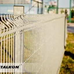 Szary, metalowy system ogrodzeniowy panelowy z widocznym logotypem producenta w lewym dolnym rogu, na tle zielonego trawnika i rozmytego boiska.