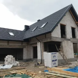 Surowy stan domu jednorodzinnego z ciemną dachówką i oknami dachowymi, widoczne materiały budowlane Solbet oraz drewniana drabina oparta o ścianę.