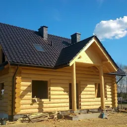 Surowy dom z bali z ciemnym dachem z blachodachówki i dwoma kominami w trakcie budowy, widoczne niezabudowane otwory okienne, jasne drewno, niebieskie niebo z chmurami w tle.