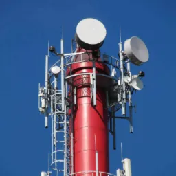 Czerwona wieża telekomunikacyjna z antenami parabolicznymi i drabinką na tle błękitnego nieba.