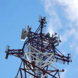 Wysoka wieża telekomunikacyjna z antenami i elementami konstrukcji stalowej, widziana z dołu na tle błękitnego nieba z lekkimi obłokami.