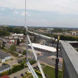 Widok z góry na miasto z elementami konstrukcji stalowej, w tym maszt antenowy i wsporniki, z widocznymi przewodami i elementami mocującymi. W tle budynki, drogi i zieleń miejska pod lekko...