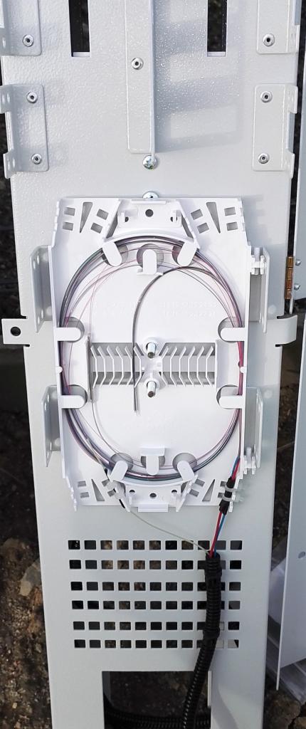 Biała skrzynka telekomunikacyjna z włóknami światłowodowymi ułożonymi w okrąg, zamontowana na metalowej konstrukcji z perforacją.