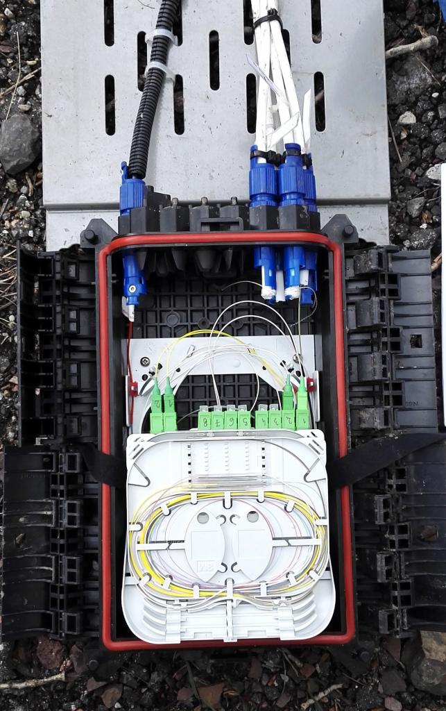 Otwarta skrzynka telekomunikacyjna z widocznymi światłowodami, złączami i modułami, umieszczona na ziemi z gruzem.