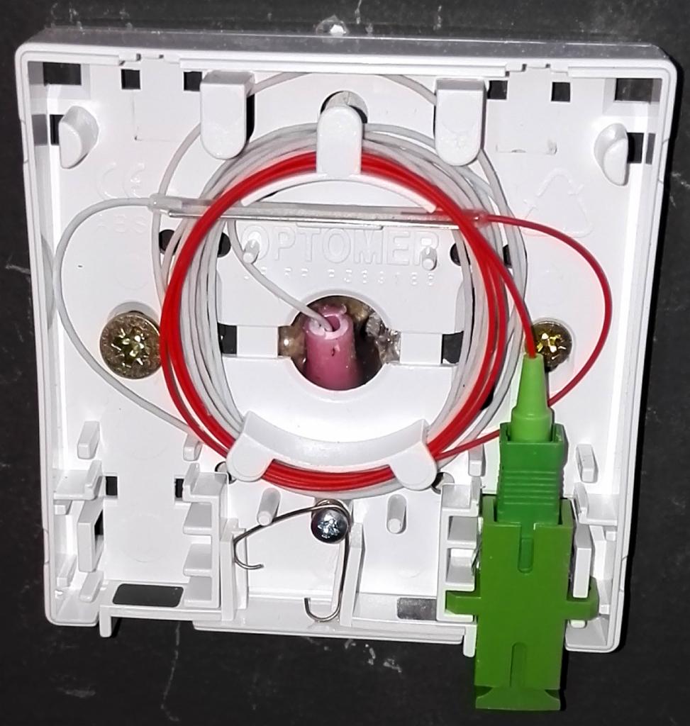 Biała puszka telekomunikacyjna z czerwonymi i białymi kablami światłowodowymi oraz zielonym złączem, widoczne śruby mocujące.