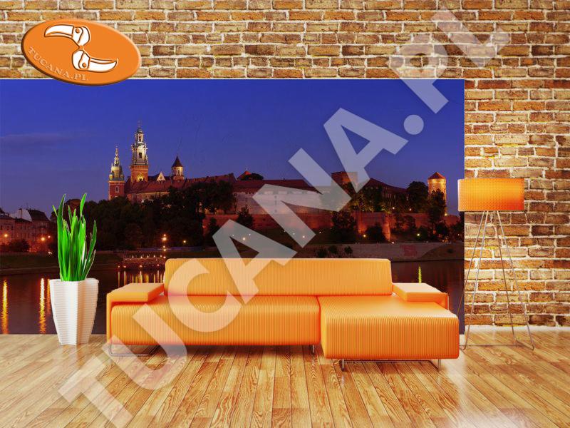 Winylowa fototapeta na fizelinie z nocnym widokiem na Wawel, umieszczona w aranżacji wnętrza z pomarańczową sofą i lampą na trójnogu.