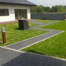 Kaskadowa ścieżka z kostki brukowej z jasną obwódką na trawniku, prowadząca od tarasu do kamiennego muru. Nowoczesny dom z ciemnym dachem w tle. Słoneczny dzień.