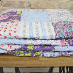 Złożona, ręcznie szyta kołdra patchworkowa w odcieniach fioletu, różu i błękitu, z motywami kwiatowymi i geometrycznymi, leżąca na drewnianym blacie z metalową podstawą w tle ceglanej ściany.