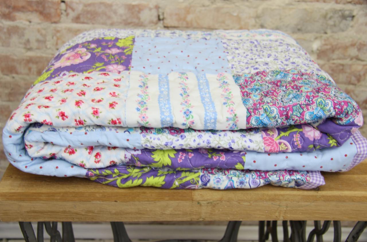 Złożona, ręcznie szyta kołdra patchworkowa w odcieniach fioletu, różu i błękitu, z motywami kwiatowymi i geometrycznymi, leżąca na drewnianym blacie z metalową podstawą w tle ceglanej ściany.