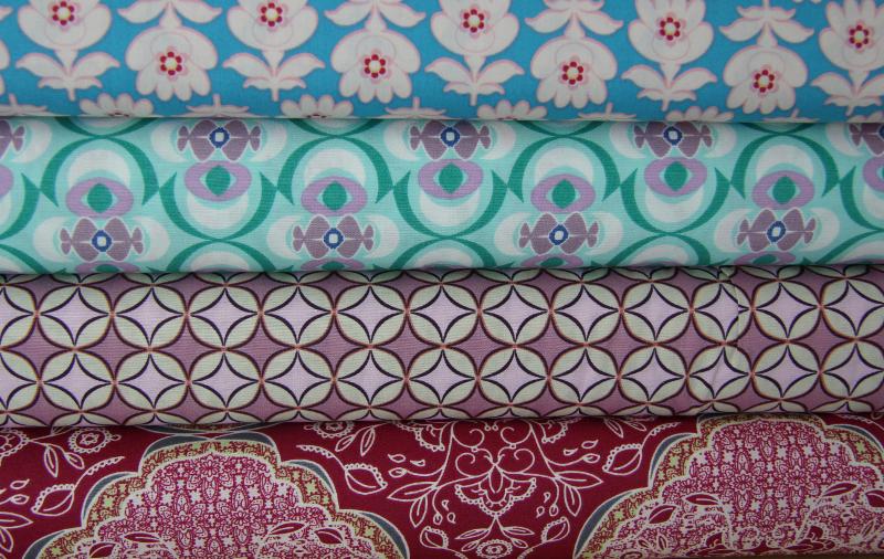 Stos tkanin bawełnianych Art Gallery Fabrics w różnych wzorach i kolorach: kwiaty na błękicie, geometryczne kształty na miętowym, romby na liliowym i paisley na czerwonym tle.