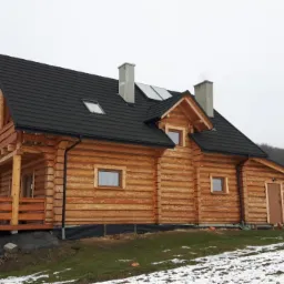 Nowo wybudowany, drewniany dom z bali z czarnym dachem i dwoma kominami, otoczony zaśnieżonym terenem, z widocznym białym samochodem dostawczym w tle.