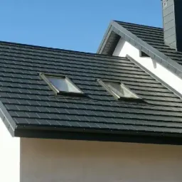 Nowoczesny dach z ciemnej dachówki ceramicznej z dwoma oknami połaciowymi, widoczny komin i orynnowanie w kolorze dachu na tle błękitnego nieba.