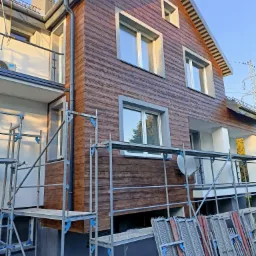 Dom z drewnopodobną elewacją w trakcie montażu, widoczne rusztowania i fragmenty elewacji do ułożenia, komin z cegły klinkierowej.