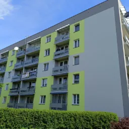 Wielopiętrowy budynek mieszkalny po renowacji elewacji, z fragmentami pomalowanymi na jasnozielony kolor i szary, balkony z szarymi balustradami, widoczne anteny satelitarne i markizy balkonowe...