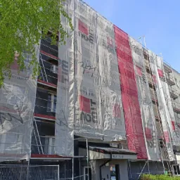 Wielopiętrowy budynek mieszkalny w trakcie prac elewacyjnych, osłonięty siatką z nadrukiem reklamowym, widoczne rusztowania i fragmenty ocieplenia ścian, zieleń drzew w lewym górnym rogu.