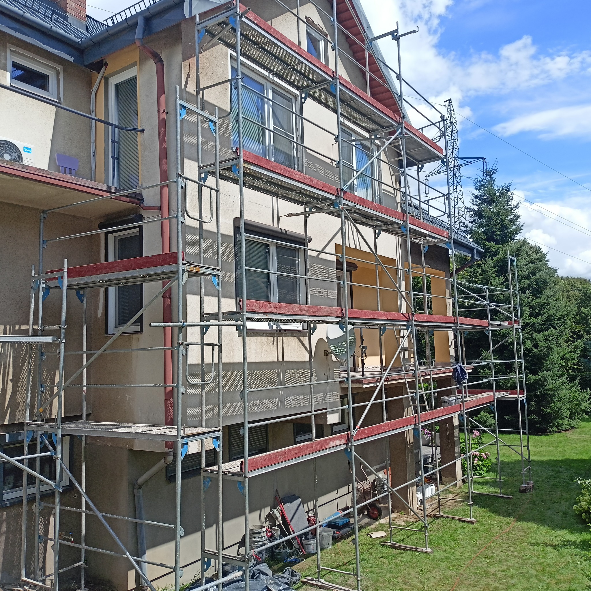 Dom jednorodzinny w trakcie prac związanych z elewacją, widoczne rusztowania, ocieplenie styropianem i przygotowane narzędzia na trawniku.