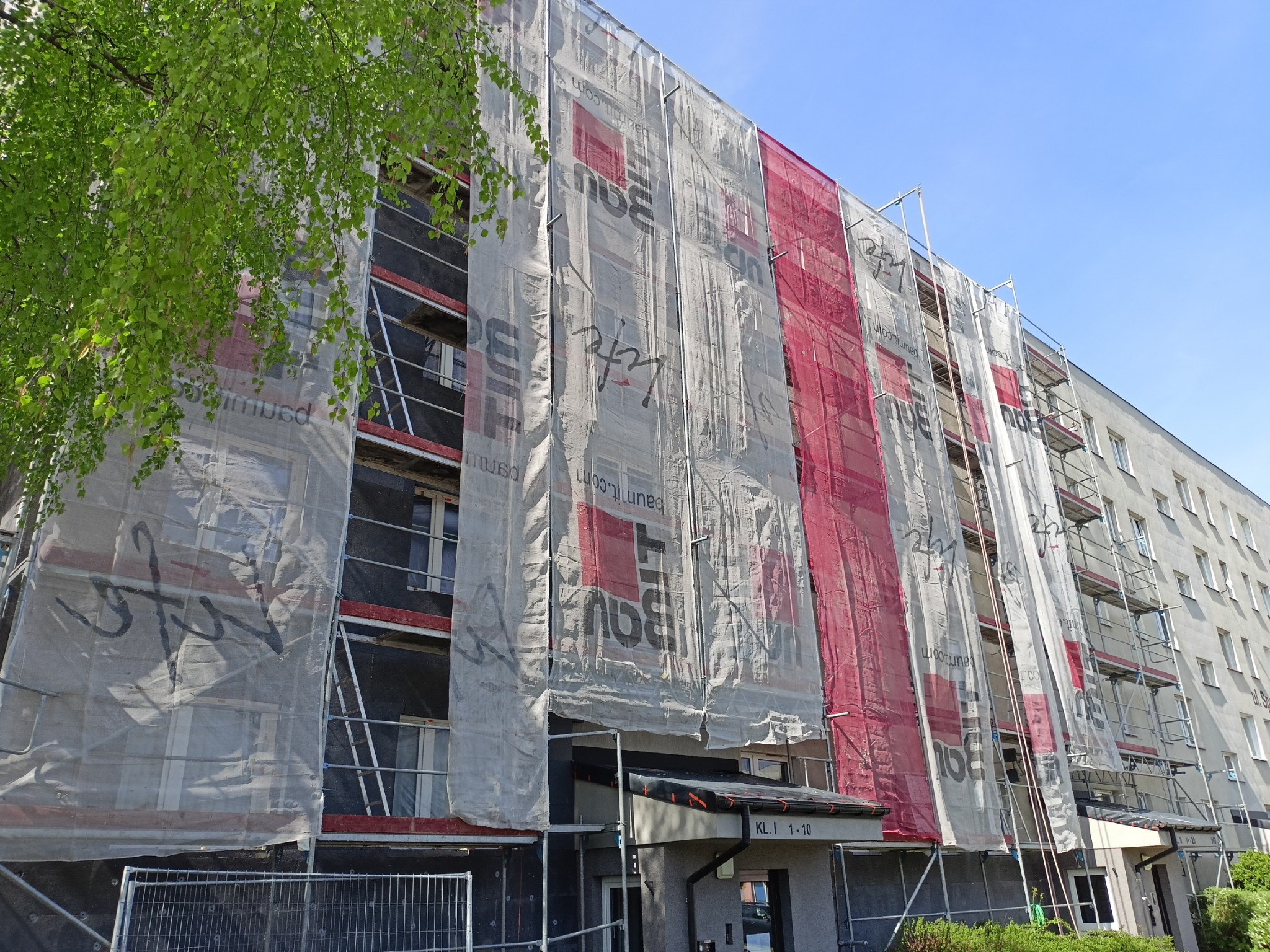 Wielopiętrowy budynek mieszkalny w trakcie prac elewacyjnych, osłonięty siatką z nadrukiem reklamowym, widoczne rusztowania i fragmenty ocieplenia ścian, zieleń drzew w lewym górnym rogu.