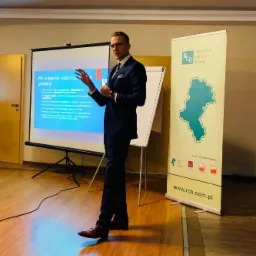 Prezentacja PPK: mężczyzna w garniturze gestykuluje przed ekranem z informacjami o Pracowniczych Planach Kapitałowych, obok roll-up Regionalnego Centrum Biznesu.