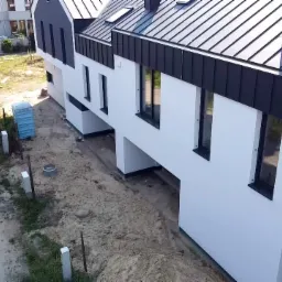 Widok z góry na szereg nowych, białych budynków z czarnymi dachami i oknami, teren wokół w trakcie prac ziemnych, w tle biegnący mężczyzna w czerwonej koszulce.