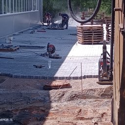 Creativ - Prace brukarskie w toku: robotnicy układają kostkę brukową obok budynku przemysłowego, widoczny wykop i maszyny budowlane, palety z kostką w tle.