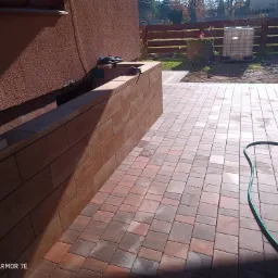 Nowo ułożona, wielobarwna kostka brukowa na posesji, widoczny fragment ściany budynku, murku, węża ogrodowego i zagęszczarki w tle.