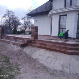 Creativ - Nowo wybudowany taras z kostki brukowej w odcieniach brązu, z niskimi murkami i schodami prowadzącymi do domu, obok stoi myjka ciśnieniowa.