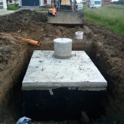 Betonowa studzienka kanalizacyjna umieszczona w wykopie, widoczna rura kanalizacyjna i koparka w tle, teren budowy przed domem.