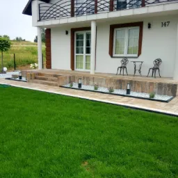 Elewacja domu z tarasem wyłożonym jasną kostką, z zielonym trawnikiem, ozdobnymi lampkami i meblami ogrodowymi w stylu retro.