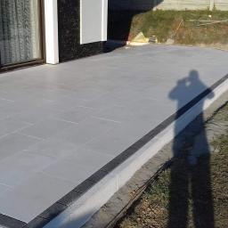 Creativ - Nowoczesny taras z szarych płyt betonowych, obrzeża z ciemnej kostki, cień fotografa widoczny na trawie, częściowo wykończony teren.