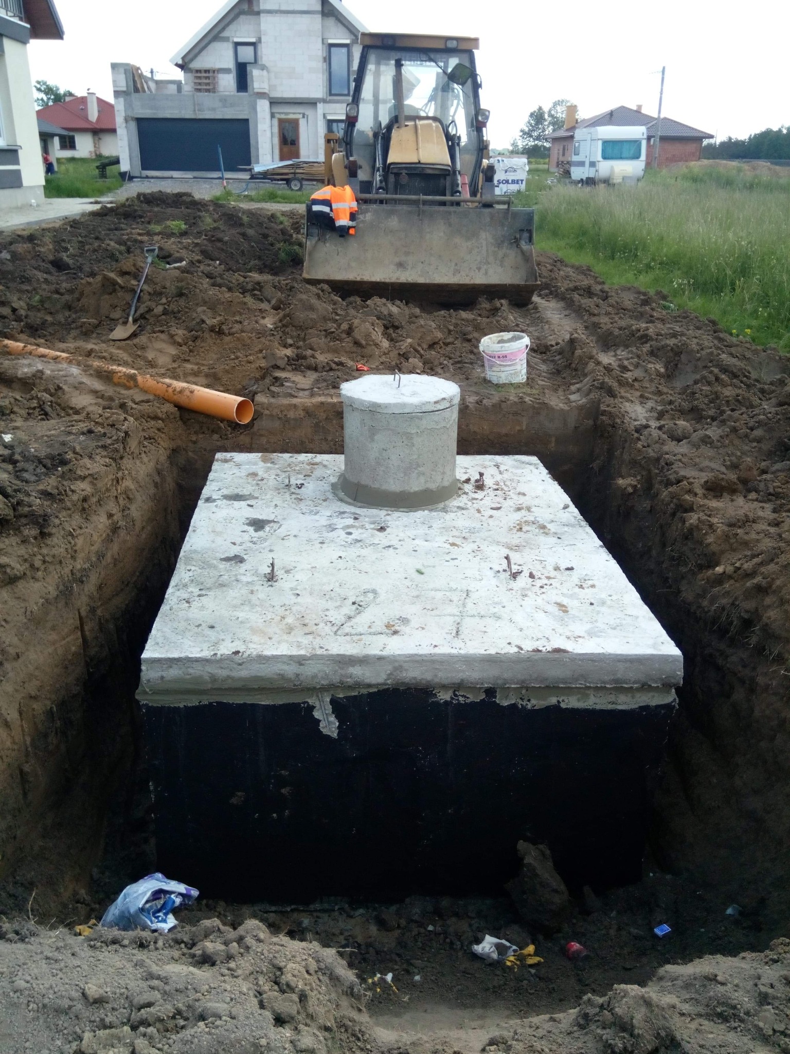 Betonowa studzienka kanalizacyjna umieszczona w wykopie, widoczna rura kanalizacyjna i koparka w tle, teren budowy przed domem.