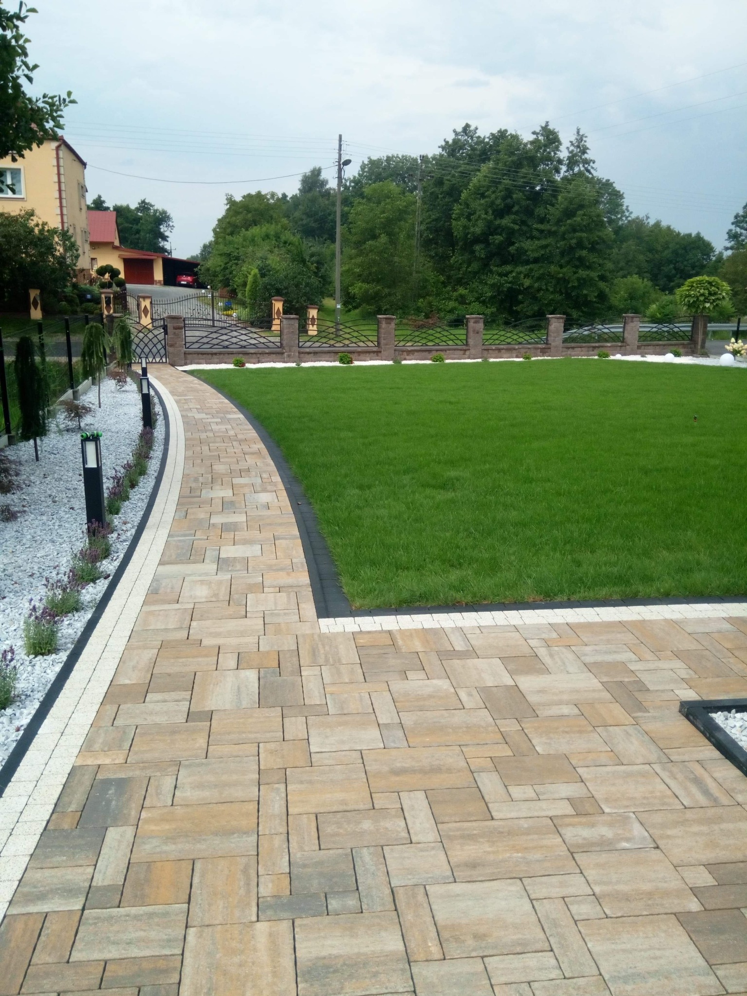 Ułożona kostka brukowa w odcieniach beżu i szarości tworzy ścieżkę przez ogród, obok trawnika i białego żwiru z niskimi lampami oświetleniowymi oraz ozdobnym ogrodzeniem z metalowymi przęsłami...