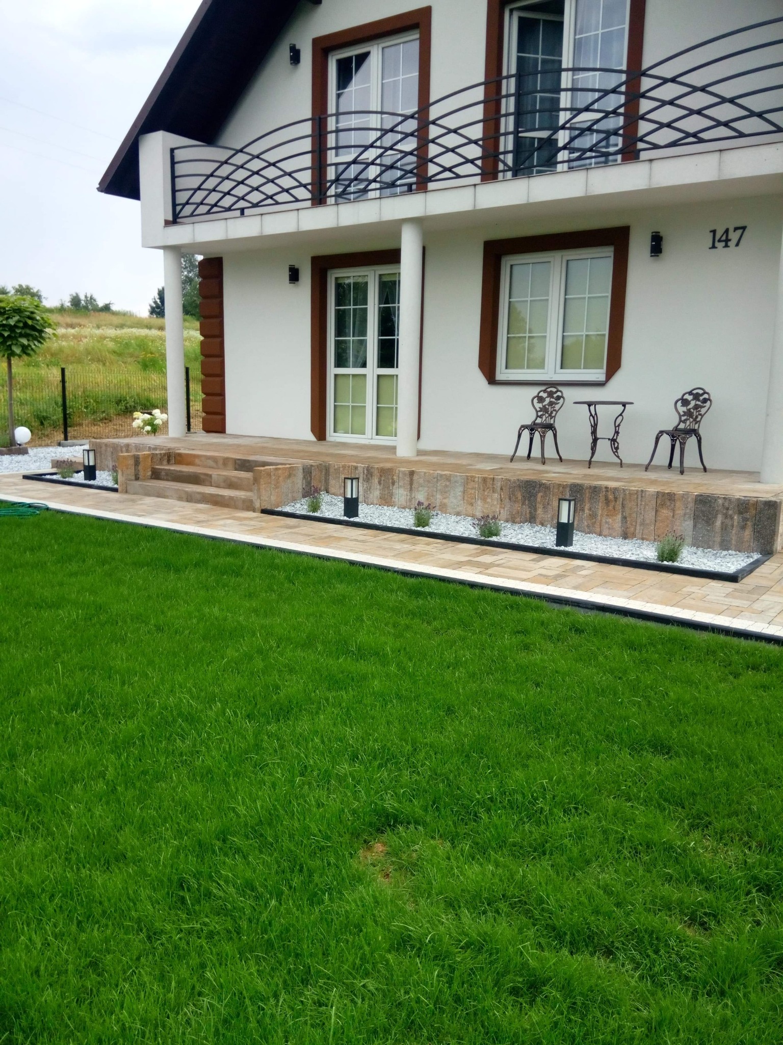 Elewacja domu z tarasem wyłożonym jasną kostką, z zielonym trawnikiem, ozdobnymi lampkami i meblami ogrodowymi w stylu retro.