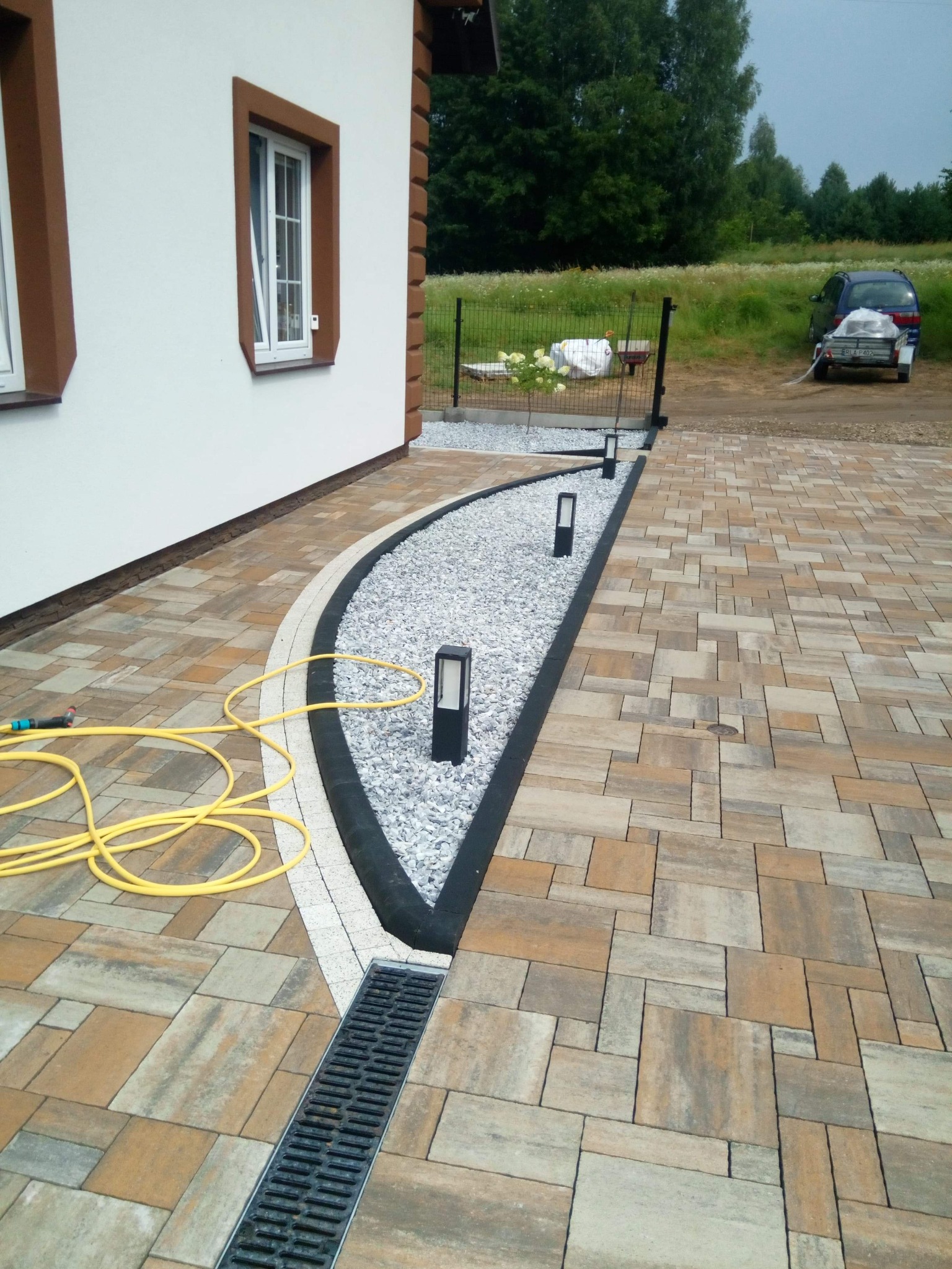 Ułożona kostka brukowa w Zalesiu z odpływem liniowym i dekoracyjnym pasem żwiru oddzielającym budynek od podjazdu, widoczne lampy ogrodowe i zwinięty wąż ogrodowy.