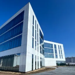 Nowoczesny budynek biurowo-laboratoryjny z elewacją szklaną wentylowaną, widziany z dołu pod ostrym kątem, z intensywnym błękitnym niebem w tle i fragmentem placu budowy u dołu kadru.