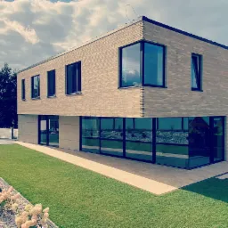 Nowoczesny dom jednorodzinny z elewacją z jasnej cegły ręcznie formowanej, duże okna z czarnymi ramami, zadbany trawnik i rabaty z białym kamieniem i kwitnącymi hortensjami.