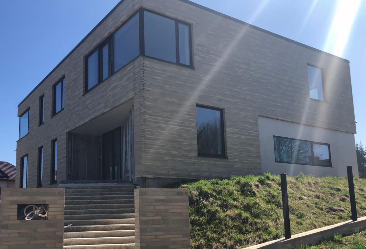 Nowoczesny dom jednorodzinny z elewacją z cegły klinkierowej w odcieniach szarości, z dużymi oknami i schodami wejściowymi z ceglanym murem ozdobionym metaloplastyką, w słoneczny dzień.