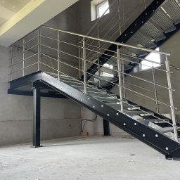 WELD-BUD - Metalowe schody z ażurowymi stopniami i balustradą ze stali nierdzewnej w surowym wnętrzu hali, widoczne okna. Element konstrukcyjny w stanie deweloperskim.