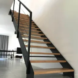 Nowoczesne schody stalowe z drewnianymi stopniami w jasnym wnętrzu. Minimalistyczny design, czarne elementy konstrukcyjne, szare płytki podłogowe, naturalne światło.