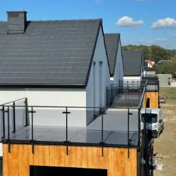 Nowoczesne domy z balkonami wykończonymi szkłem i drewnem, białe ściany, grafitowe dachy, błękitne niebo. Widoczne samochody dostawcze i osoba w oddali.