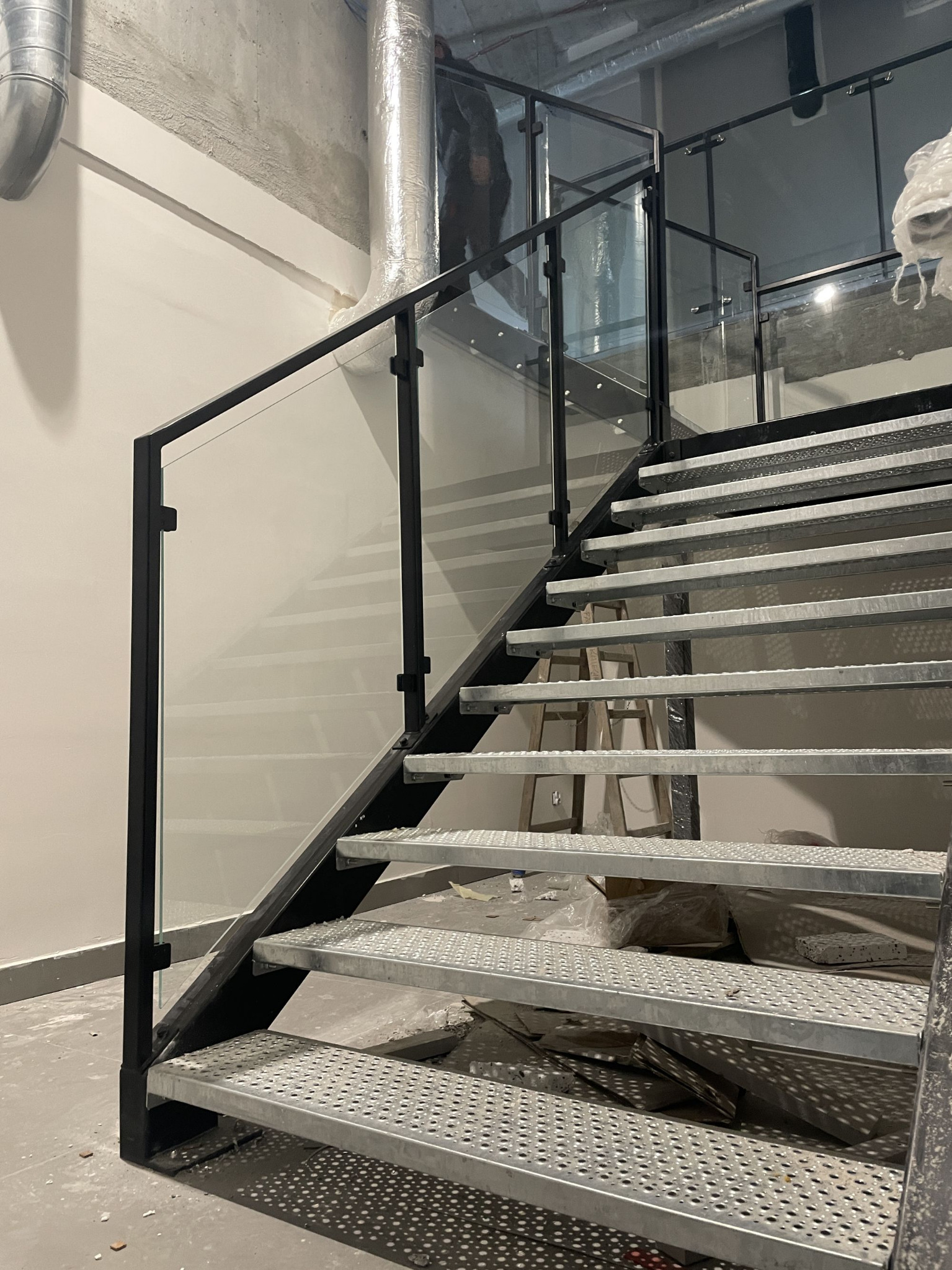 Metalowe schody z czarną balustradą i szklanym wypełnieniem w surowym wnętrzu. Widoczne elementy instalacji wentylacyjnej i drabina w tle.