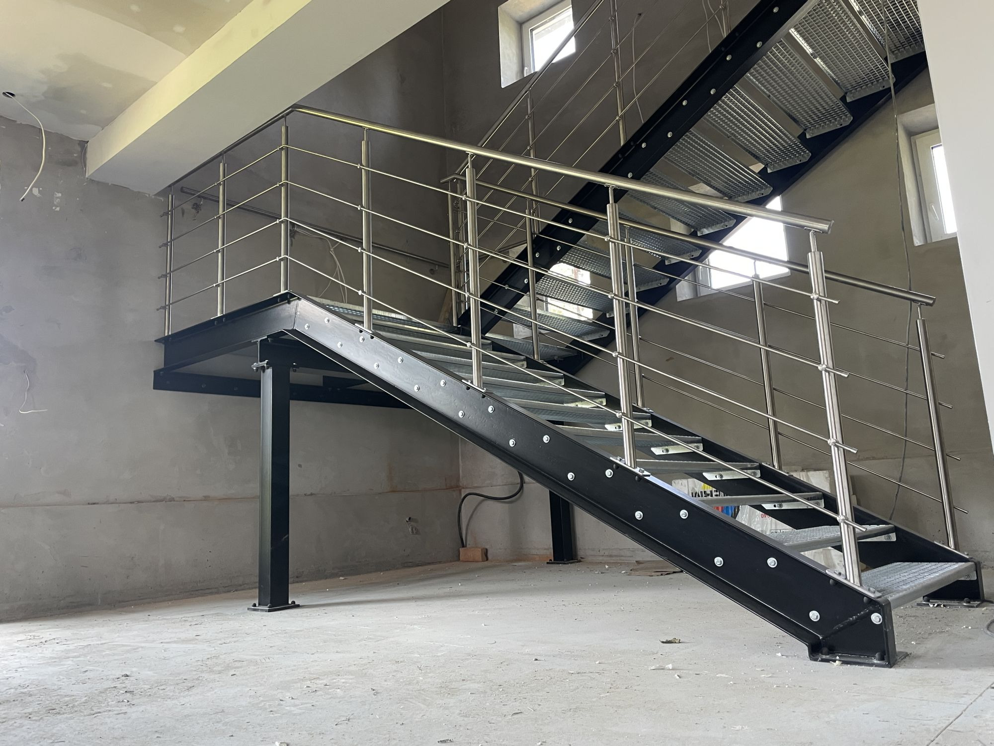 Metalowe schody z ażurowymi stopniami i balustradą ze stali nierdzewnej w surowym wnętrzu hali, widoczne okna. Element konstrukcyjny w stanie deweloperskim.