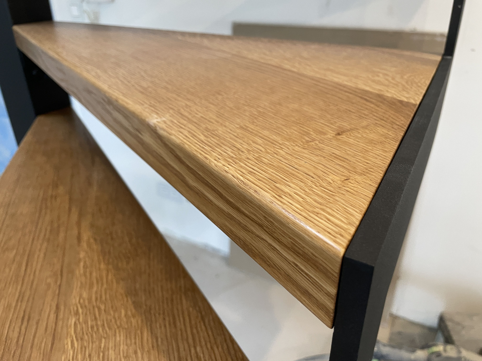 Drewniane schody z czarną metalową konstrukcją, widok z dołu, ukazujący teksturę drewna i precyzję wykonania. Minimalistyczny design, jasne oświetlenie.