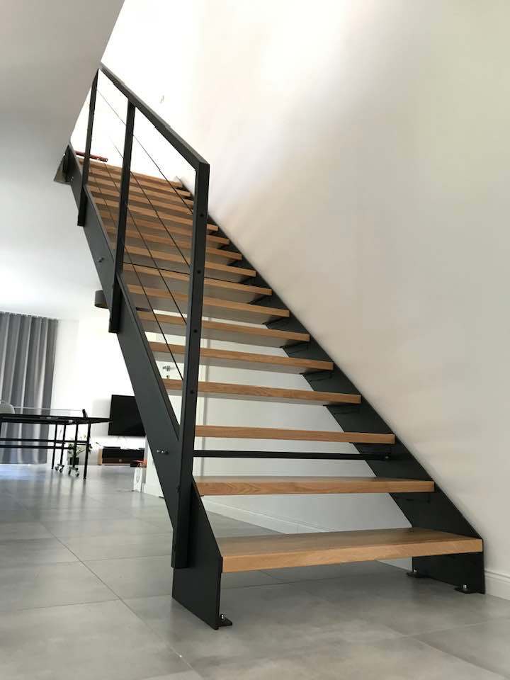 Nowoczesne schody stalowe z drewnianymi stopniami w jasnym wnętrzu. Minimalistyczny design, czarne elementy konstrukcyjne, szare płytki podłogowe, naturalne światło.