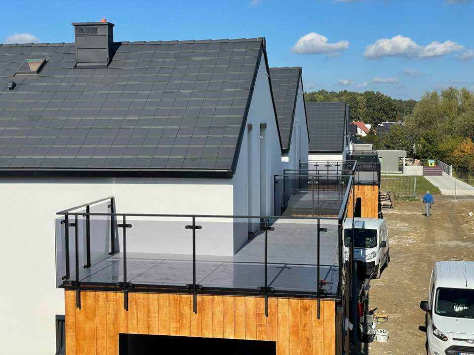 Nowoczesne domy z balkonami wykończonymi szkłem i drewnem, białe ściany, grafitowe dachy, błękitne niebo. Widoczne samochody dostawcze i osoba w oddali.